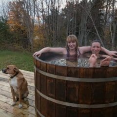 Two women in cedar hot tub and a dog outside the tub.